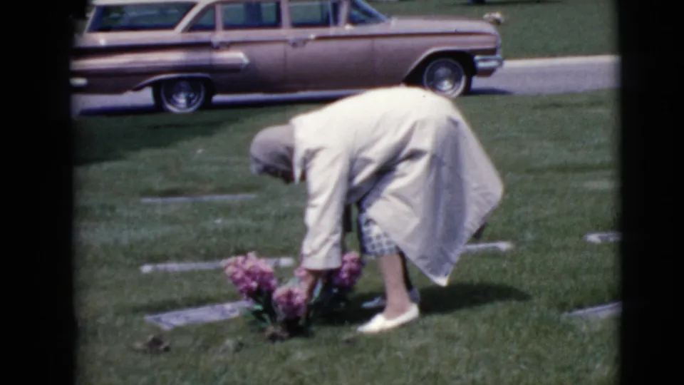 In the cemetery, the woman leans low over a cluster of pink flowers placed beside flat grave markers on the grass.