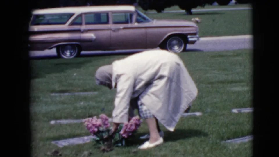 The 1968 home-movie frame shows a woman in a light coat tending pink flowers beside a grave marker in Clarksdale, Arizona.