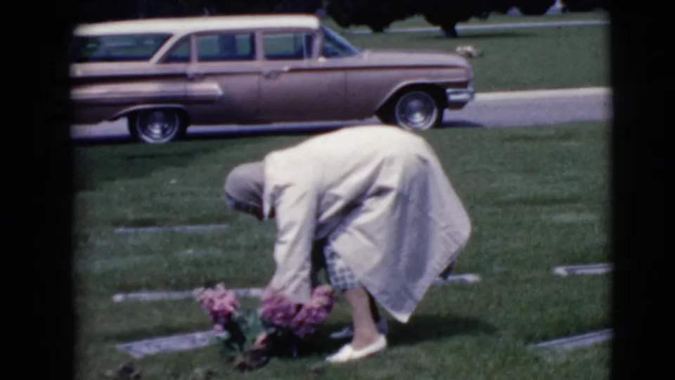 In metadata-listed Clarksdale, Arizona, 1968, a woman in a light coat bends over a grave with pink flowers as a station wagon sits behind her.