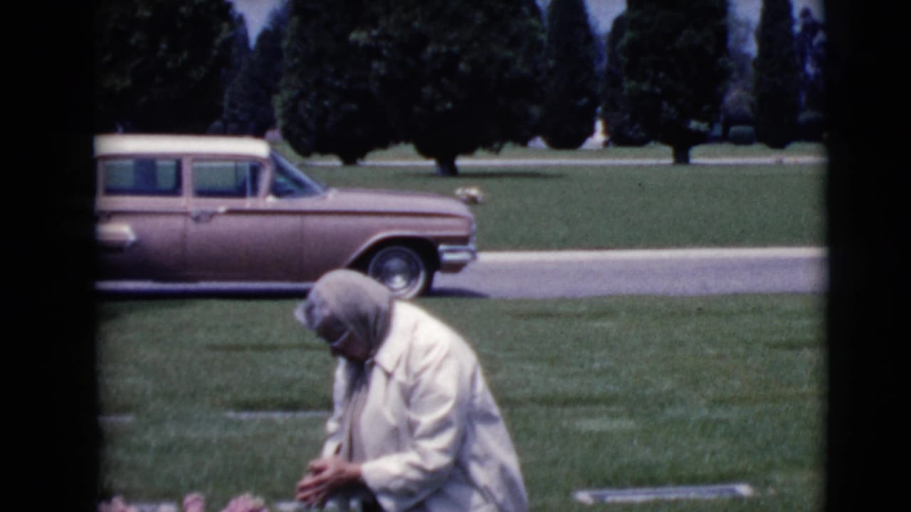 Woman kneels or crouches near a grave with a station wagon parked behind her.
