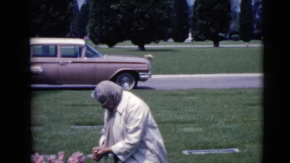 The woman continues tending the grave in a sunlit cemetery, with pink flowers near the foreground and the parked 1960s wagon behind her.