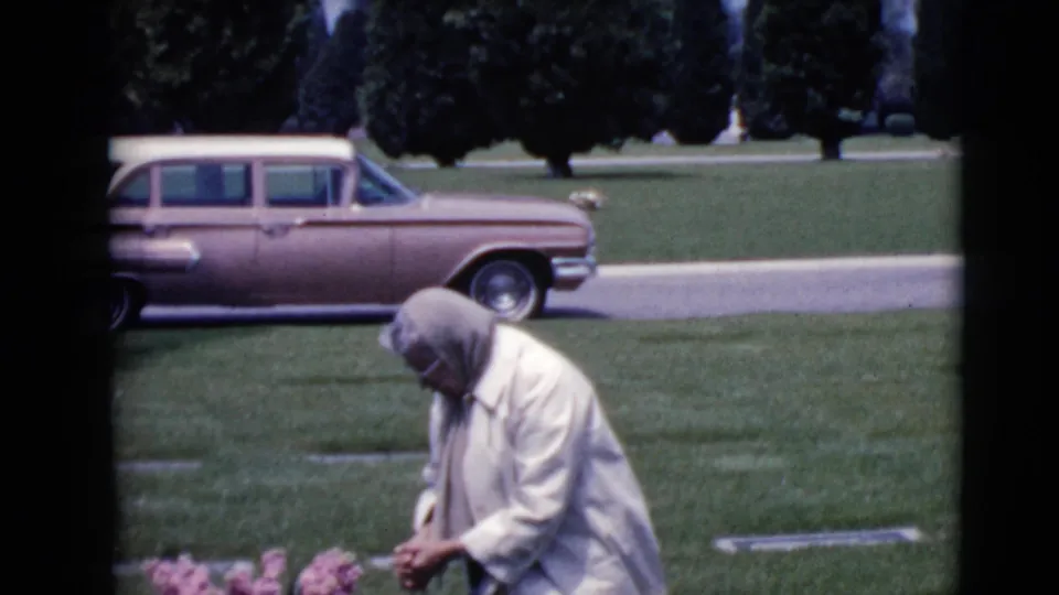 At the end of the clip, the woman appears to pause with hands clasped near the grave, framed by lawn, markers, road, trees, and the station wagon.