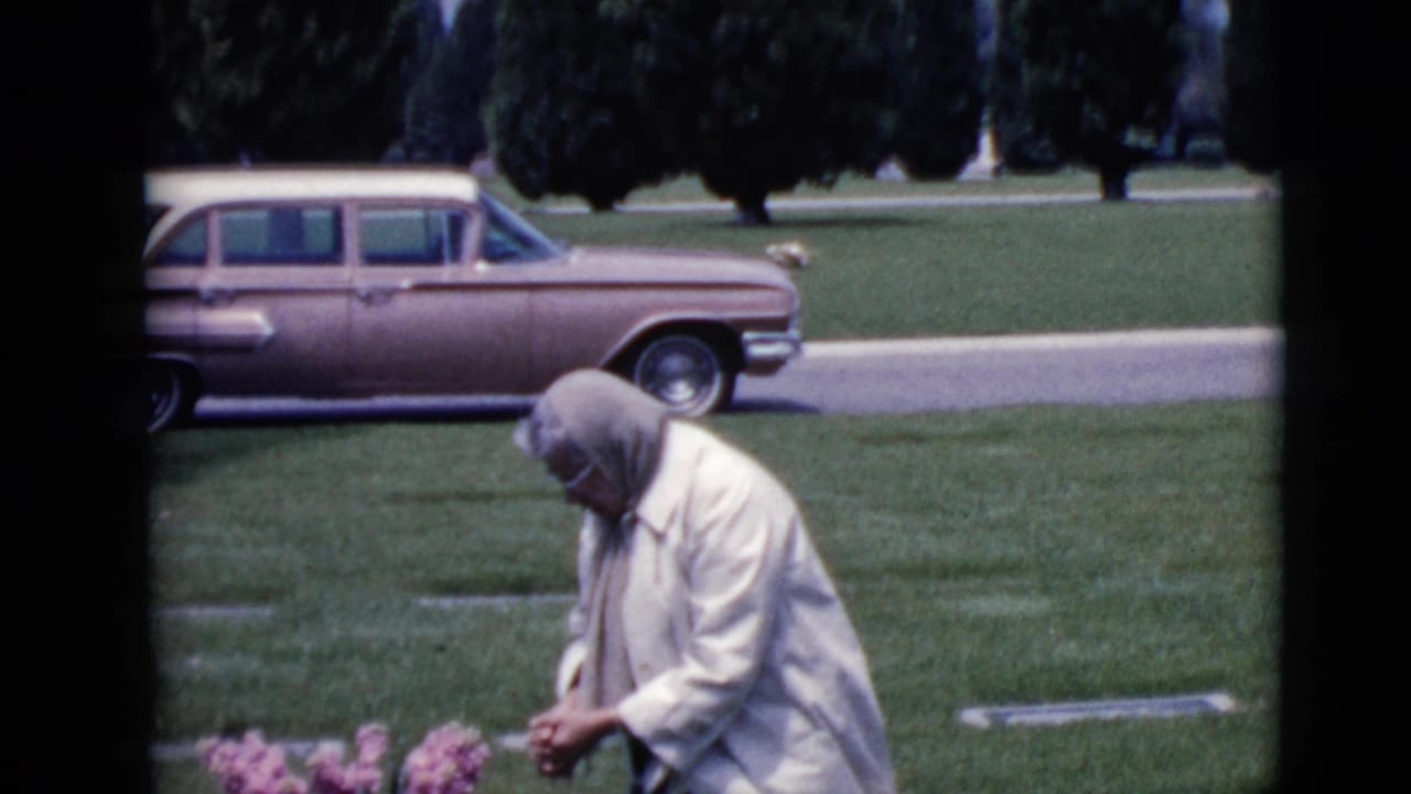 Woman in a light coat and headscarf bends near pink flowers in a cemetery lawn.