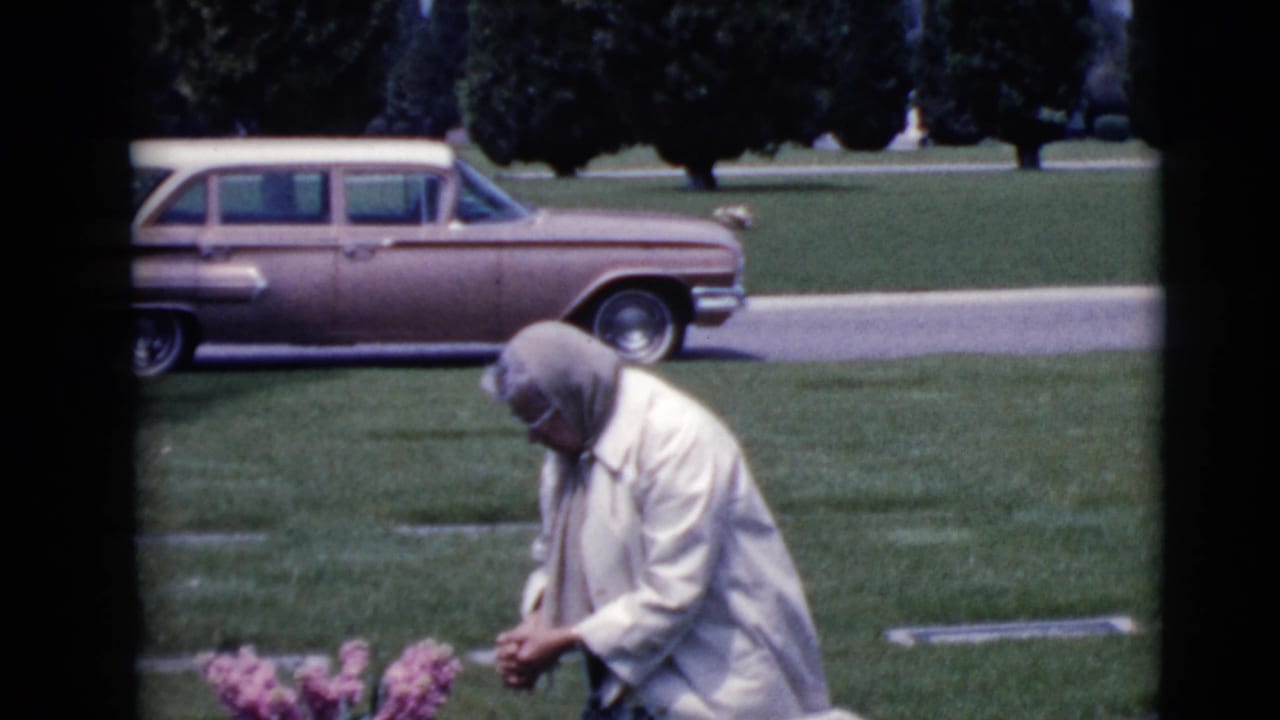 Woman kneels at a grave site with pink flowers and a station wagon behind her.