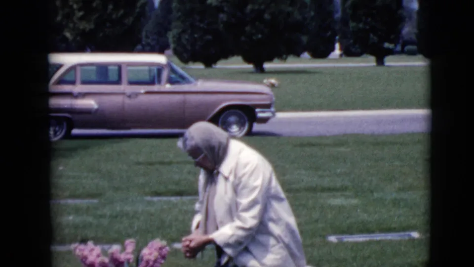 A 1968 home-movie frame shows the woman leaning toward graveside flowers in a Clarksdale cemetery, with a period station wagon in the background.