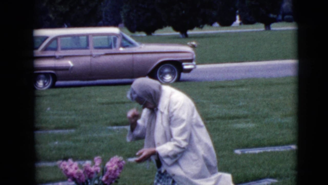 Elderly woman kneels by pink flowers in a cemetery with a station wagon behind her.