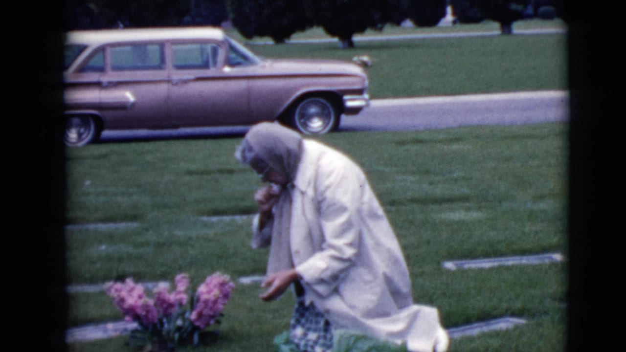 The woman remains bent over beside pink flowers in the cemetery, with flat markers and a 1960s station wagon visible behind her.