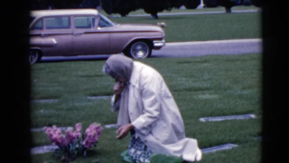 An elderly woman in a headscarf kneels low by pink flowers at a grave, with cemetery grass and a vintage station wagon behind her.
