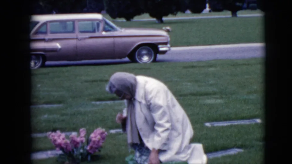 In the final frame, the woman bows over the grave area near pink flowers, with Clarksdale cemetery lawn and a parked wagon in view.