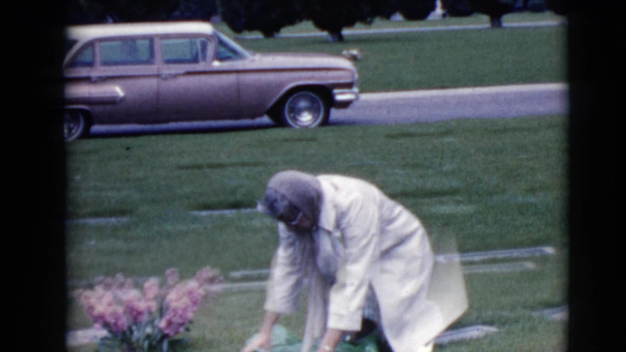 Elderly woman bends near pink flowers in a grassy cemetery with a station wagon behind her.