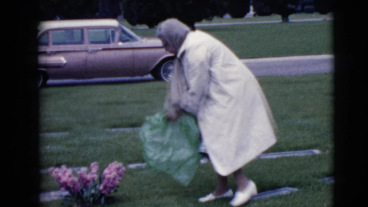 Woman stands beside a grave holding green wrapping near pink flowers and a parked station wagon.