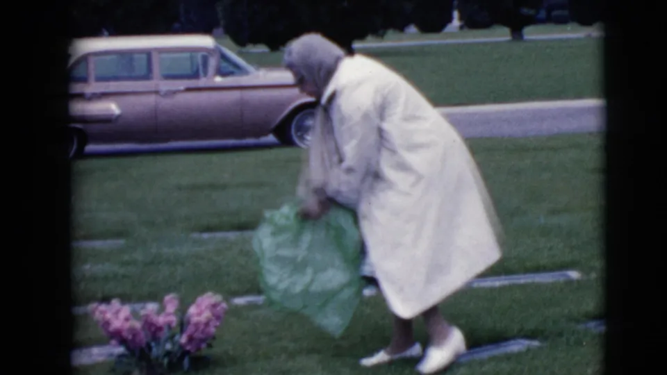 The woman stands at the cemetery grave with green flower wrapping, with pink flowers and a 1960s station wagon visible behind her.