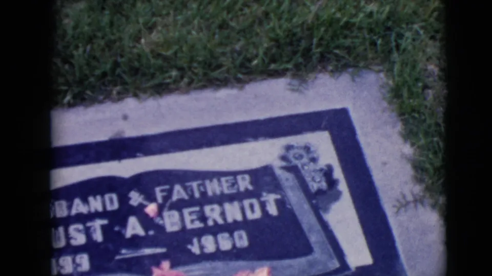 Partial handheld 1968 frame of the Berndt grave marker, cropped at the left, with grass and flowers visible.