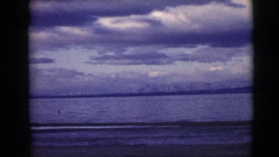 1968 8mm frame of a calm water horizon under heavy clouds, with dark frame borders and faded blue-purple color.