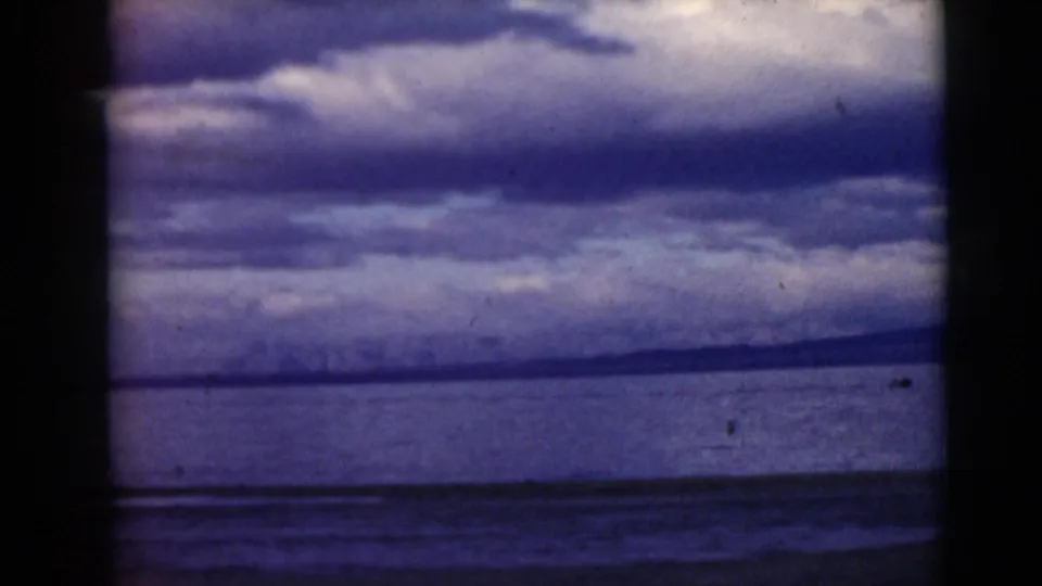 1968 8mm frame with distant shoreline, dark water bands, and a heavy cloud bank rendered in blue-purple faded color.