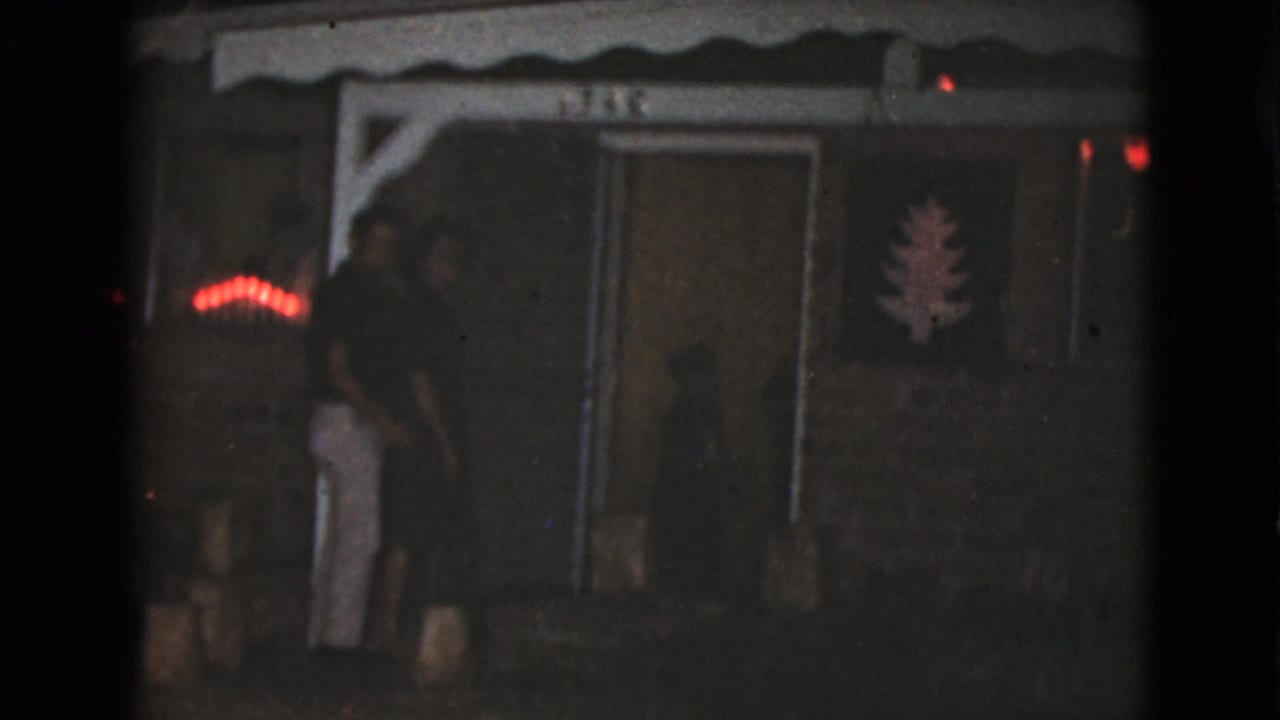 Two adults stand on a dim porch beside red lights and a tree decoration.