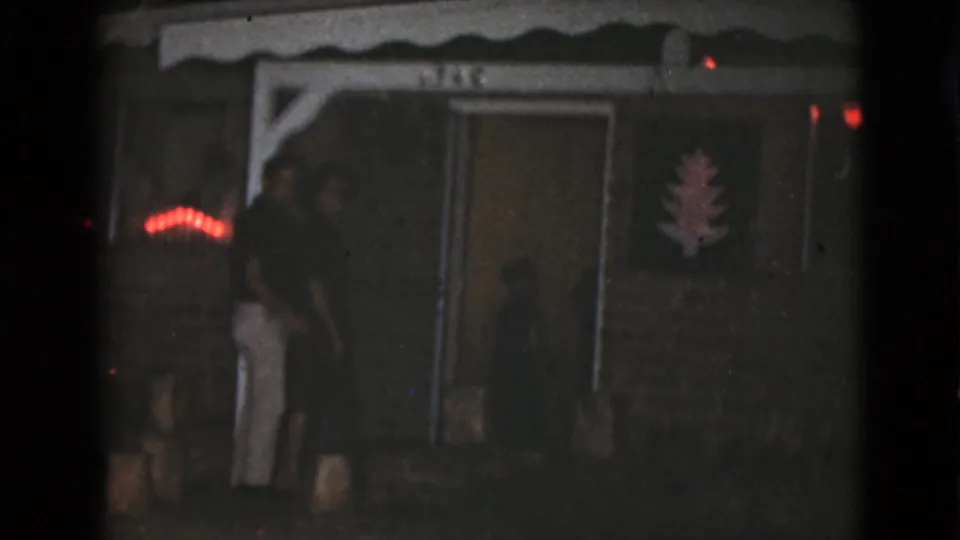 In 1968 Clarksdale, Arizona home-movie footage, two adults stand near a modest porch entrance with red lights and a wall tree decoration.