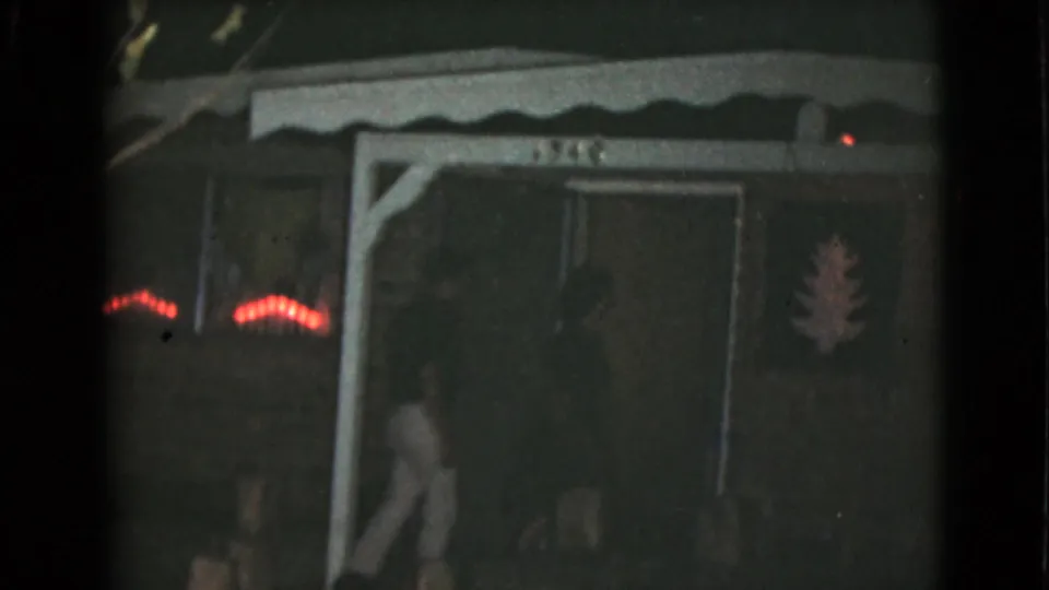 One adult steps along the porch toward the entry while another figure is partly visible near the door in the 1968 home-movie frame.