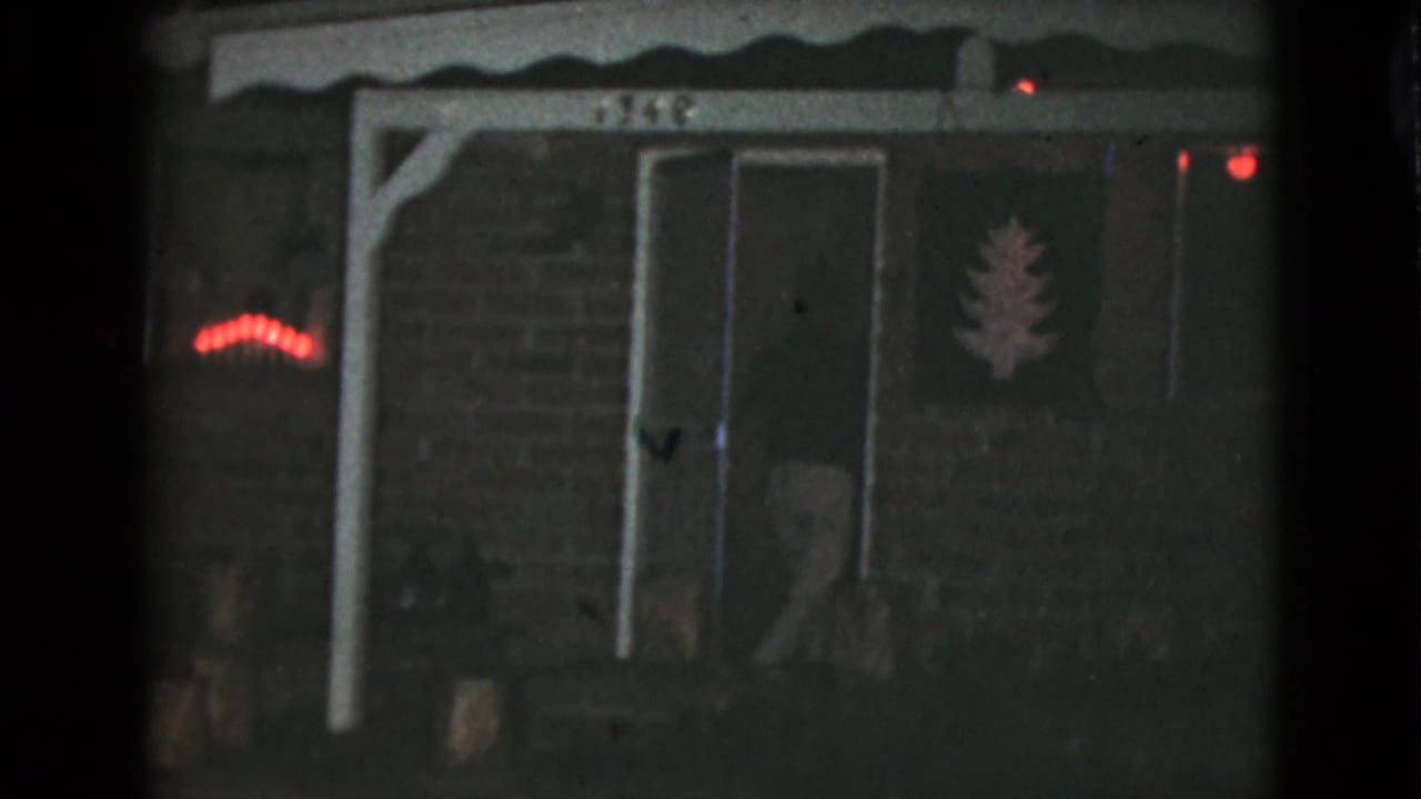 Dark brick house entrance with porch posts, red lights, and a faint person near the doorway.