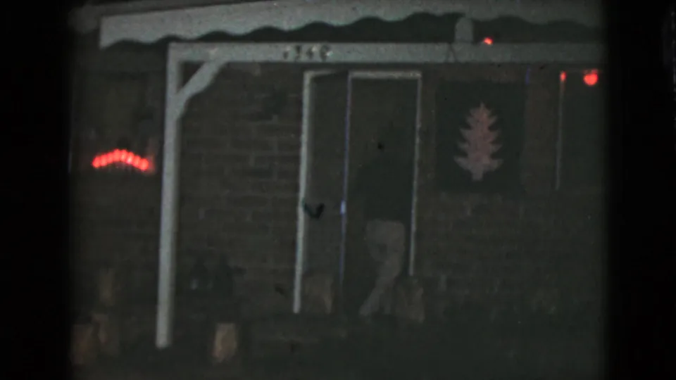 1968 8mm frame of a Clarksdale, Arizona house entrance at night, with red lights and a faint figure by the doorway.