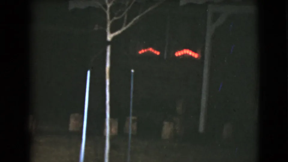 Nighttime 8mm frame from the Clarksdale, Arizona clip showing dark trees, vertical posts, and faint red lighting in the distance.