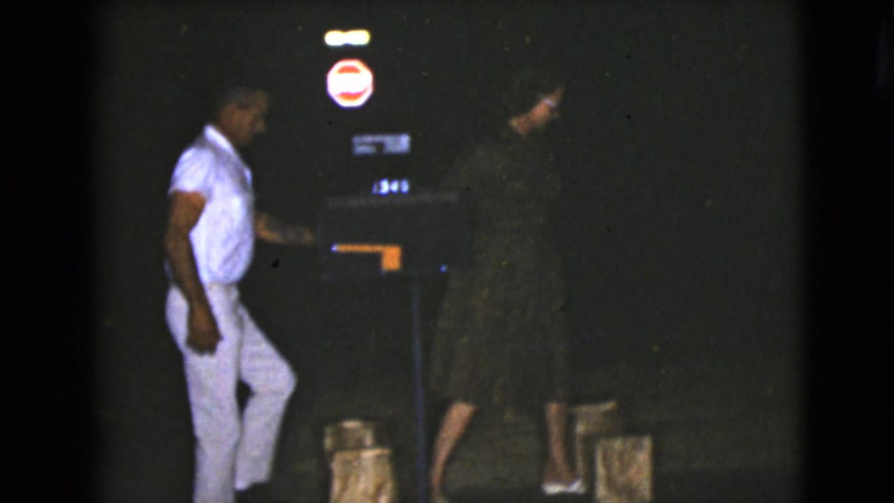 In the 1968 Clarksdale, Arizona night scene, a man in light clothing faces a woman in a dark dress near illuminated signage.