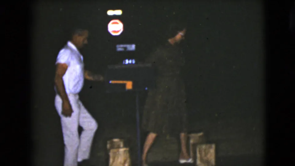 In the 1968 Clarksdale, Arizona night scene, a man in light clothing faces a woman in a dark dress near illuminated signage.
