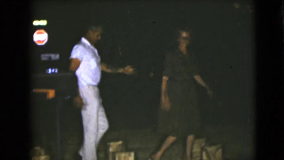 The 8mm frame shows the adult pair near nighttime signage, with the man extending a hand toward the woman in dark clothing.