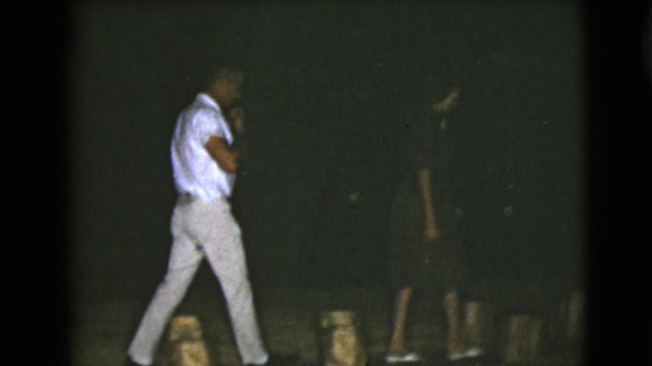 Two men walk at night across a dark outdoor area with stump-like blocks near the ground.