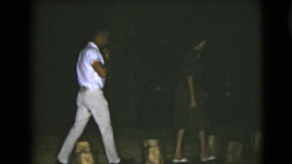 In 1968 Clarksdale, Arizona home-movie footage, two adult men walk through a dim outdoor area, one in light clothing and one in dark clothing.