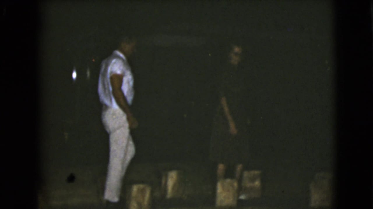 A man in a white shirt and light trousers appears in dim 1968 Clarksdale, Arizona outdoor footage, with a darker figure partly visible in the background.