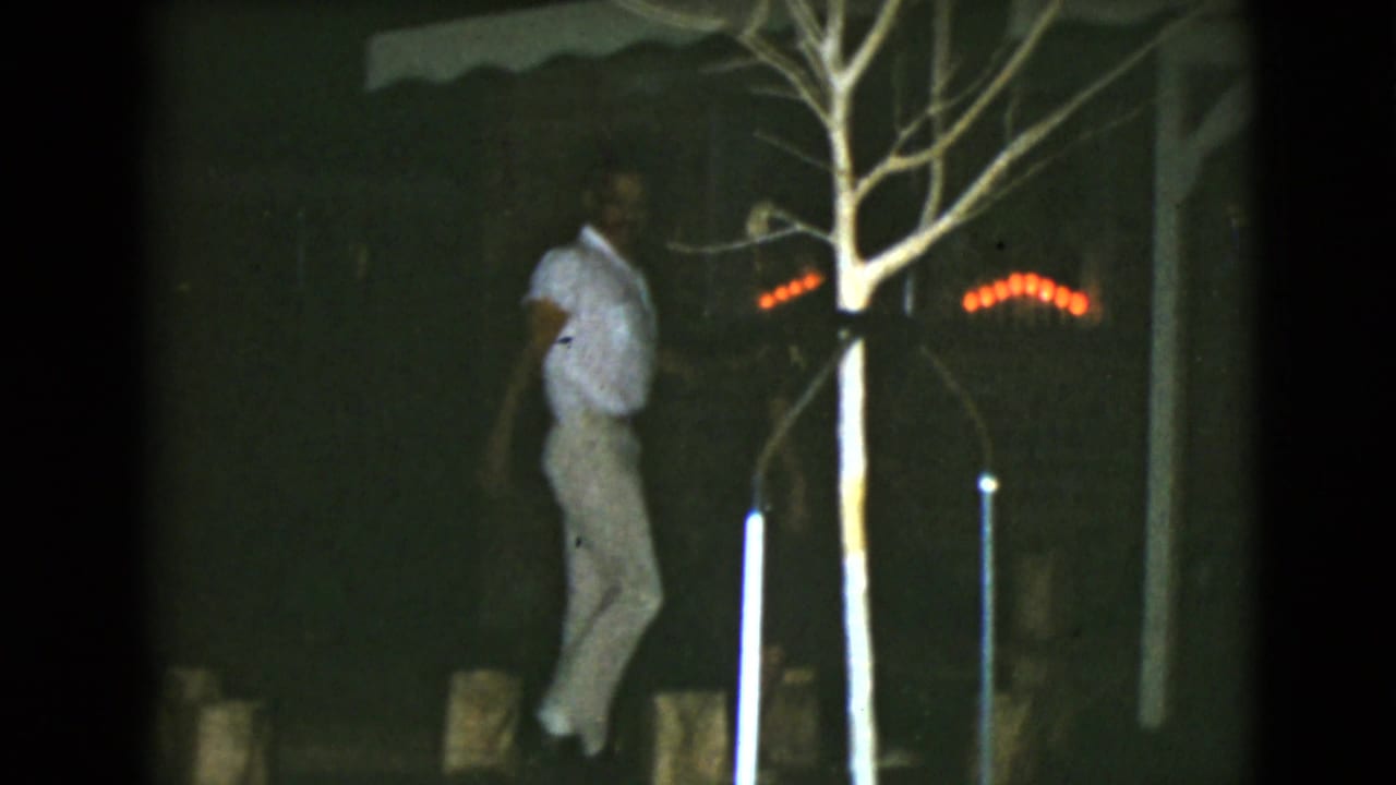 Man in light clothing walks past a bare tree toward a brick house at night.