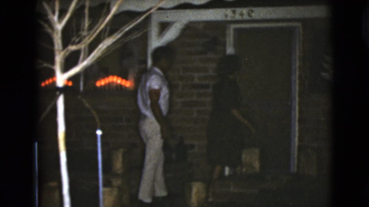 Man stands near the front door of a brick house with red lights behind him.