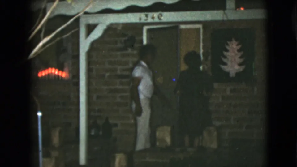 A man in light clothing and a woman in a dark dress stand by the front door of a Clarksdale brick house at night.