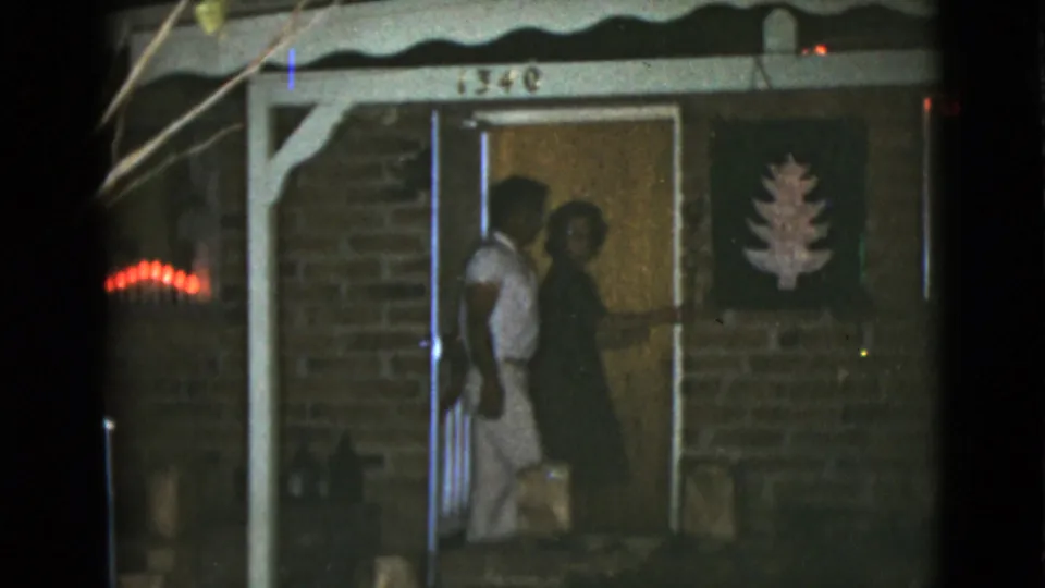 The couple is framed at the open doorway, with porch posts, brickwork, and a tree-shaped decoration visible in the night scene.