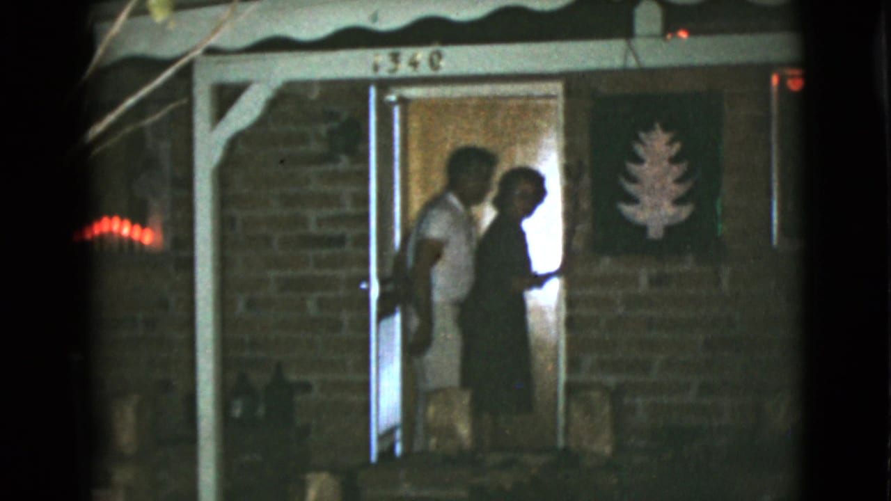 Two adults stand in a lit doorway of a brick house at night.