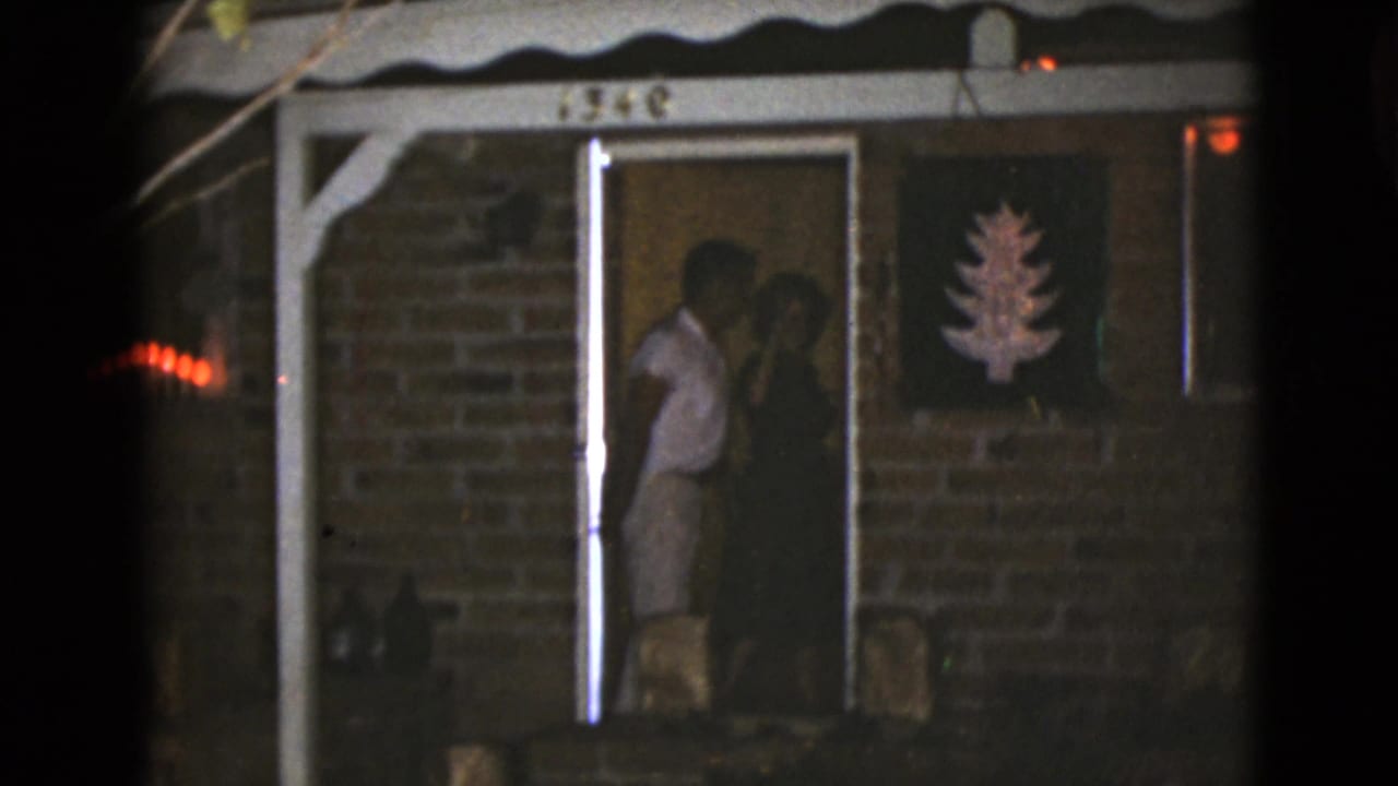 Two adults stand partly inside the lit doorway of the brick house, with the tree decoration visible on the wall beside them.