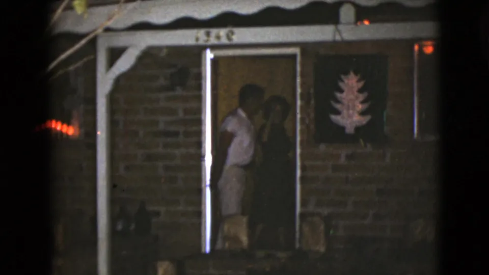 Two adults stand partly inside the lit doorway of the brick house, with the tree decoration visible on the wall beside them.
