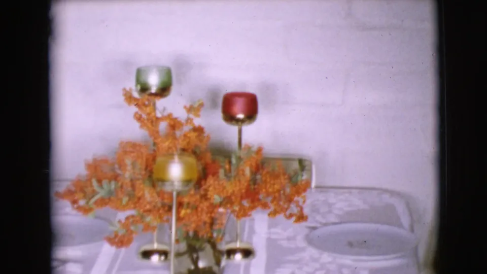 1968 home-movie interior showing a holiday dining table with orange floral arrangement, gold candle holders, plates, and a pale patterned tablecloth.