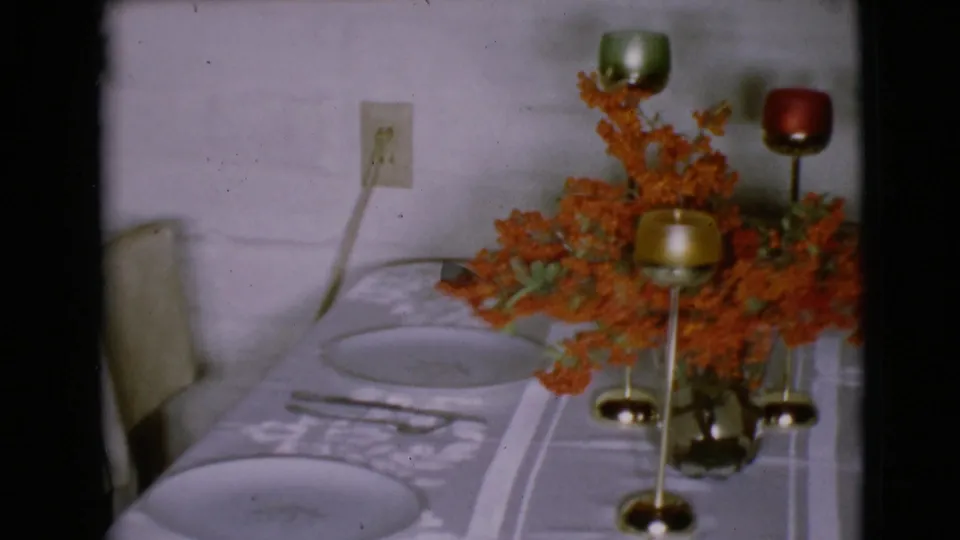 1968 interior frame showing the holiday table from the side, with orange flowers, colored candle cups, plates, a chair, and a wall outlet visible.