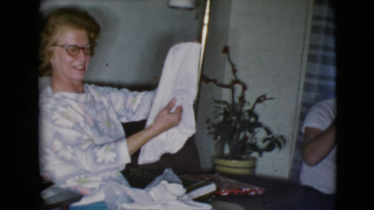 The woman smiles in the private home interior as she lifts the white cloth present, with a plant and striped curtain behind her.