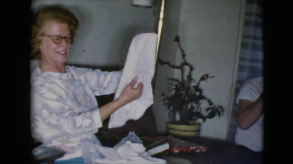 The woman smiles in the private home interior as she lifts the white cloth present, with a plant and striped curtain behind her.