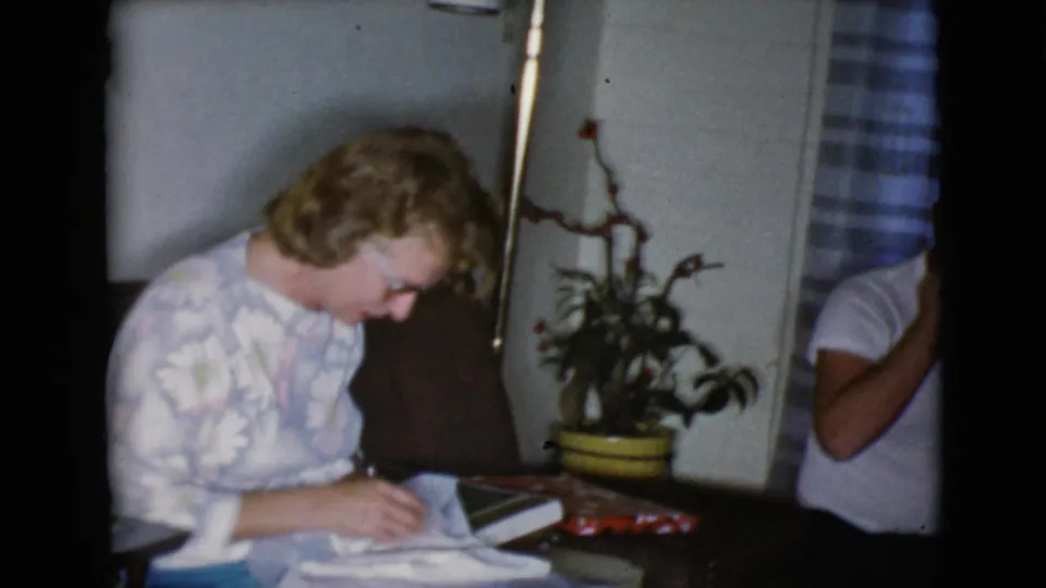 The woman leans forward over the white cloth gift on her lap in a softly lit 8mm home interior.