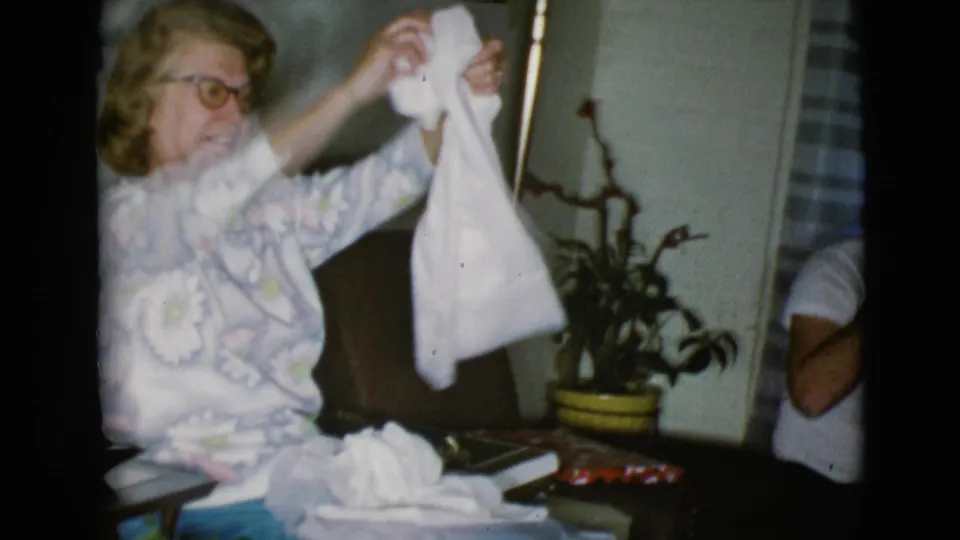 The woman raises the white cloth gift with both hands, smiling in the 1968 Arizona home-movie scene.