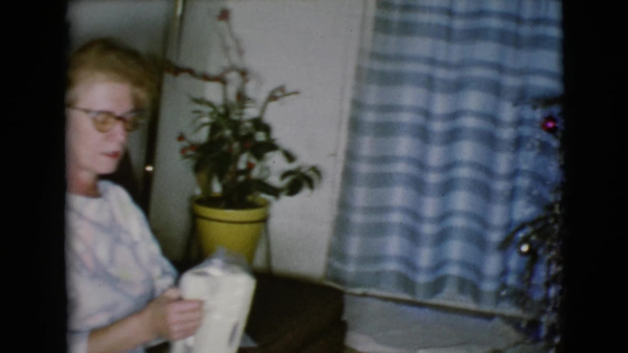 Woman in glasses sits indoors holding a white wrapped or boxed object beside a potted plant and striped curtain.