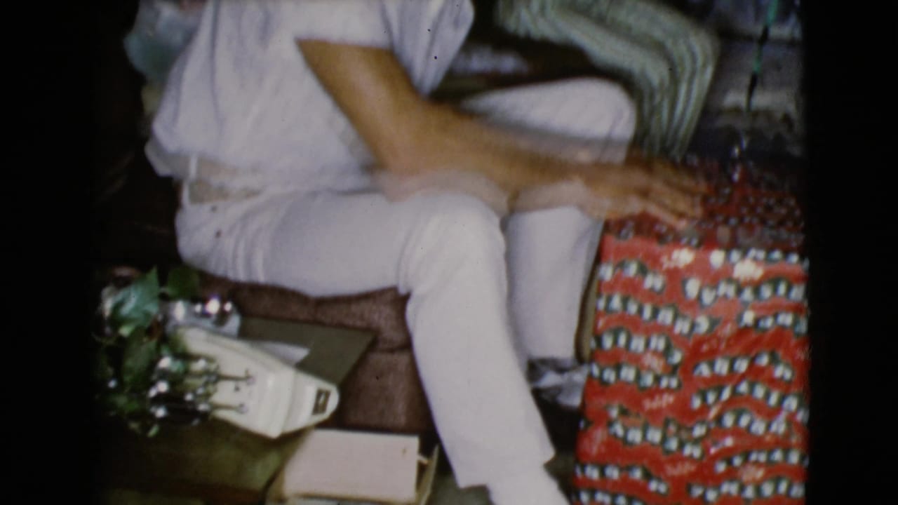 A seated adult in white clothing reaches toward red patterned wrapping paper in a 1968 Clarksdale, Arizona Christmas home interior.
