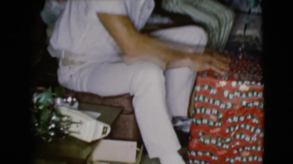 A seated adult in white clothing reaches toward red patterned wrapping paper in a 1968 Clarksdale, Arizona Christmas home interior.