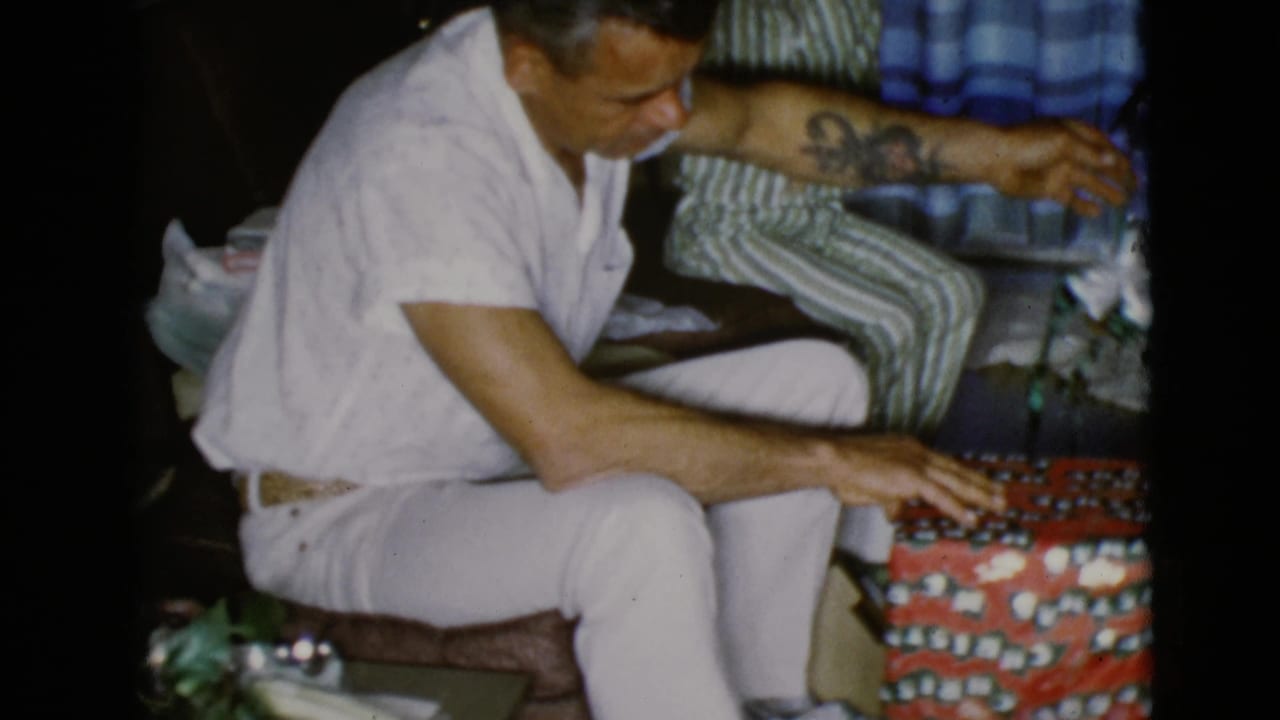 Man in white clothing sits on a couch with his hand on a red patterned gift as a child sits behind him.