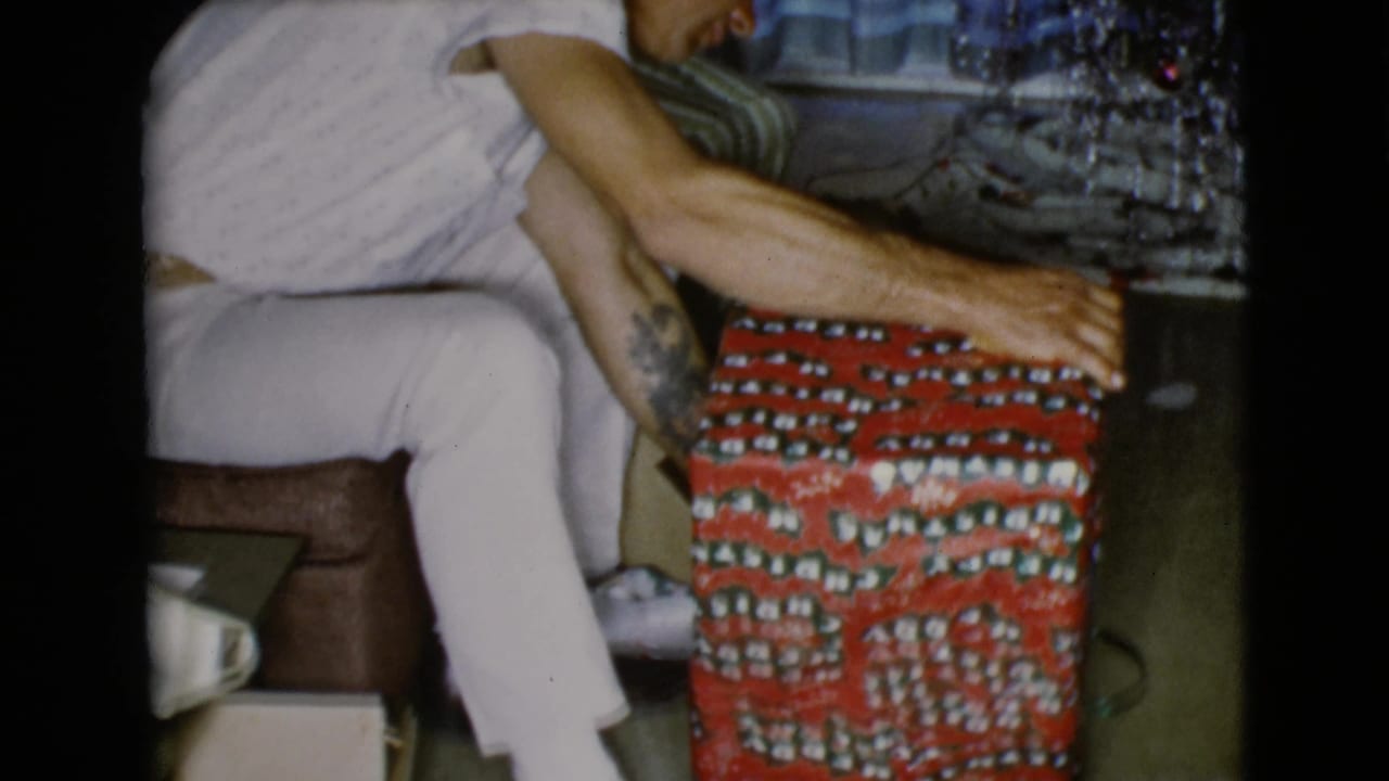 Close side view of the seated man reaching toward a red patterned gift inside a dim room.