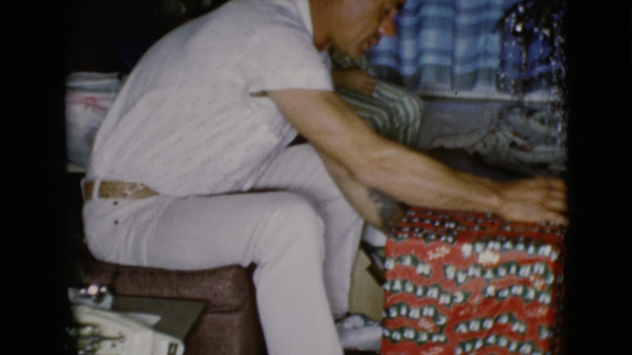 In the home interior, the man bends toward the wrapped gift as a child in striped pajamas sits behind him near blue checkered curtains.